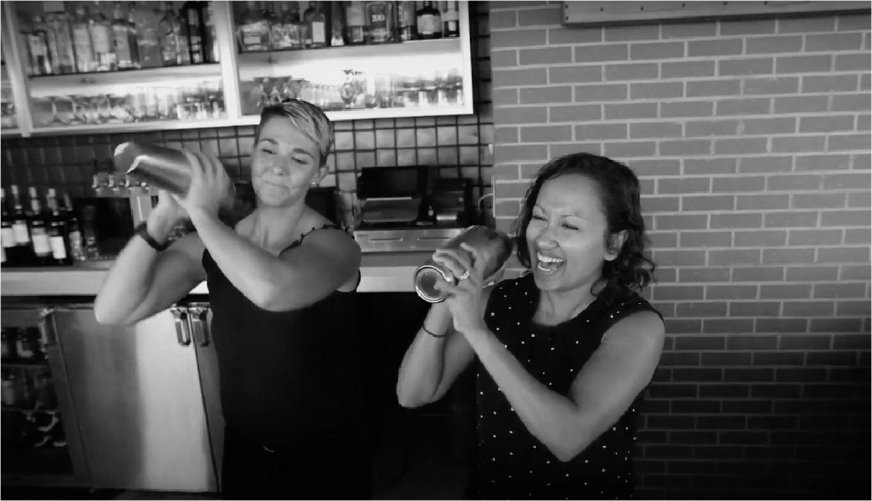 Two women shaking drink tumblers behind the bar and smiling.