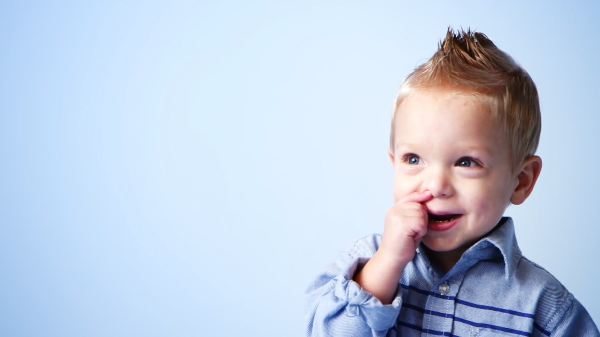 A little boy with a blonde mohawk laughing while picking his nose.