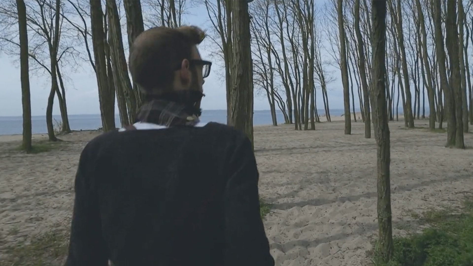 Man with a scarf and sweater is walking through a forest of bear trees near the beach.