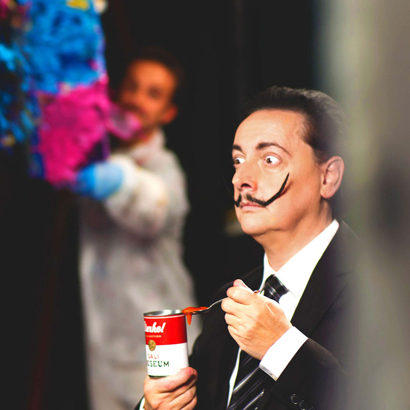 A man dressed as Salivdor Dali holding an andy Warhol soup can is about to be covered in incoming blue, pink, and yellow paint.